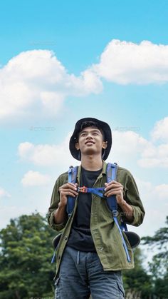 a young man wearing a hat and backpack standing in the grass with trees behind him