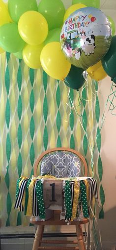 a baby's highchair in front of a birthday backdrop with green and yellow balloons