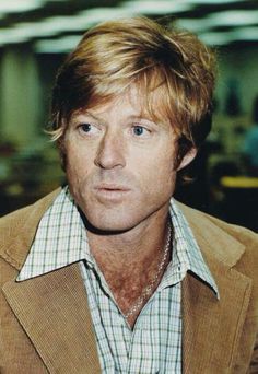 a man with blonde hair wearing a brown jacket and checkered shirt looking at the camera