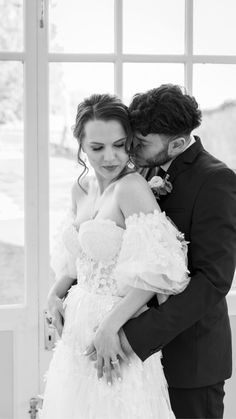 Model couple embracing in wedding attire in front of French doors of country Manor.  Bride wears a layered white lace bustier and tulle upper-arm cuffs, embellished with lace elements and with a voluminous layered tulle and lace skirt.  Groom wears dark suit.  Black and white photo
Model poses in front of an arbour, decorated with blue, blush and deep purple floral arrangements to match her bouquet.  She wears a blush and ivory multi-layered tulle bridal dress, with lace elements on bodice. Instagram Direct, Hair Accessories Jewelry