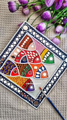 an artistic tile is laying on the ground next to purple flowers and a paintbrush