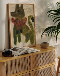 a wooden dresser topped with a book and a painting next to a potted plant