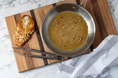 a wooden cutting board topped with a metal pan filled with food next to a piece of bread