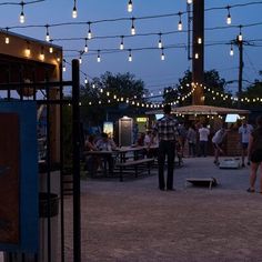 people are gathered around tables with lights strung over them at dusk in an open area