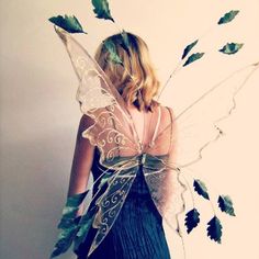 a woman dressed as a fairy with green leaves on her back and wings, standing in front of a white wall