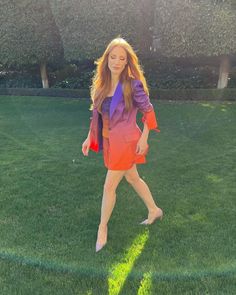 a woman in an orange and purple dress is walking across the grass with her hand on her hip