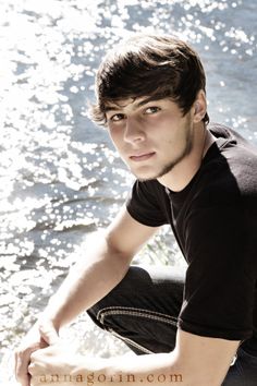 a young man sitting by the water with his hands on his knees looking at the camera