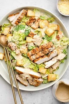 a white plate topped with chicken and lettuce next to two gold spoons