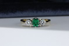 an emerald and diamond three stone ring sitting on top of a white cloth covered table