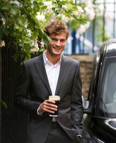 a man in a suit is holding a drink and standing next to a black car