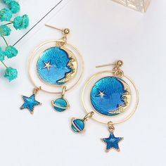 "Beautifully designed earrings featuring a moon and celestial theme with lapis-blue colors. The earrings are drop/dangle style hook. They will drop down 3\". One half of the set features a Saturn-like planet charm within an open metal circle, and a star at the bottom. The planet charm measures approximately 0.5\" in diameter, and the width of the star is approximately 0.3\". The other half of the set features a star at the top, then a round charm near the base. The round charm measures approxima Blue Dangle Hoop Earrings, Blue Metal Dangle Hoop Earrings, Blue Drop Earrings With Dangling Charms, Blue Round Earrings With Moon Charm, Blue Moon-shaped Metal Jewelry, Blue Dangle Earrings With Moon Charm, Celestial Style Drop Earrings With Dangling Charms, Blue Moon Shaped Earrings With Moon Charm, Blue Crescent Moon Charm Earrings