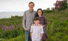 two adults and a child standing in front of a house on a hill near the ocean