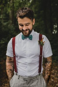 a man with tattoos wearing suspenders and a bow tie smiles at the camera while standing in the woods