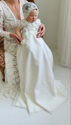 a woman holding a baby wearing a white gown and headpiece with lace on it