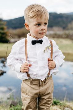 a young boy wearing a bow tie and suspenders standing in front of a lake