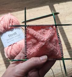 a hand holding yarn and knitting needles in front of a pink piece of cloth on a wooden surface