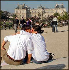 two people sitting on the ground in front of a large building with lots of people standing around