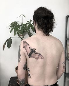 a man with a bat tattoo on his back and shoulder, standing next to a potted plant