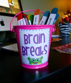 a pink bucket filled with markers and pencils on top of a desk next to a computer