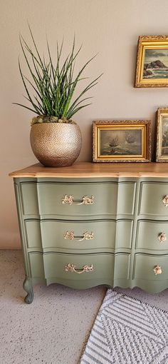 a green dresser with drawers and pictures on the wall next to potted plant in gold vase