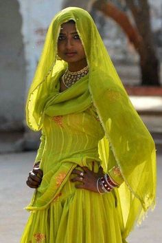 a woman in a yellow dress and head scarf is standing on the street with her hands behind her back