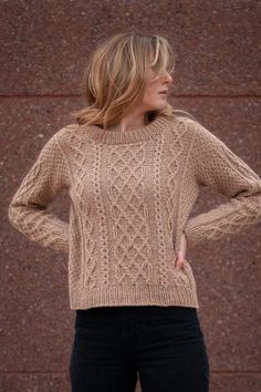 a woman standing in front of a brick wall wearing a brown sweater and black pants