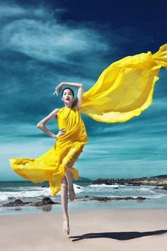 a woman in a yellow dress is running on the beach with her arms spread out