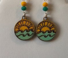 Large and long wooden circles, etched with a sun an sea design. The sea is painted an aqua green and the sun a golden yellow. Each drop is topped by two small faceted glass beads with similar color accents. Bohemian Yellow Earrings For Beach, Hand Painted Yellow Earrings For Summer, Blue Artsy Jewelry For Beach, Yellow Beach Jewelry For Vacation, Yellow Beach Vacation Jewelry, Yellow Beach Season Jewelry For Vacation, Yellow Jewelry For Beach Vacation, Beach Wooden Bead Earrings, Yellow Round Earrings For Beach