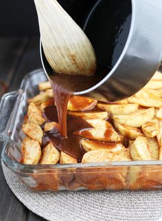 someone pouring sauce on some french fries in a glass dish with a wooden spatula