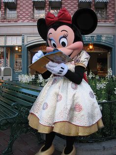 the minnie mouse mascot is reading a book in front of a park bench and building