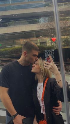 a man and woman taking a selfie in front of a glass wall with buildings behind them