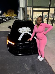 a woman in pink standing next to a black car with white bows on the hood