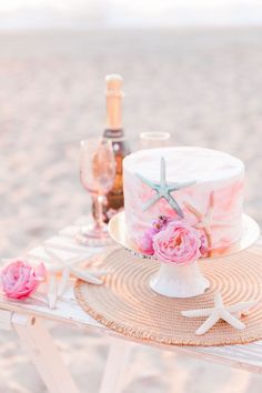 there is a cake on the beach with pink flowers and starfishs next to it