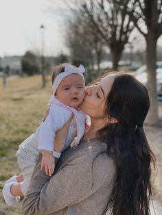 a woman is holding a baby and kissing her face in front of a park with cars