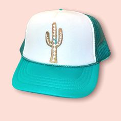 a turquoise and white hat with a gold cactus embroidered on the front, against a pink background