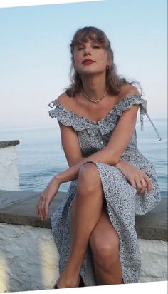 a woman sitting on the edge of a wall next to the ocean with her legs crossed