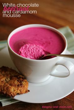 a white cup filled with pink food next to a cookie on top of a plate