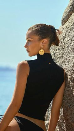 a woman leaning against a rock with her back to the camera and wearing gold earrings