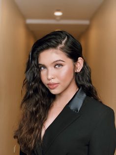 a woman with long dark hair wearing a black suit and posing for the camera in an empty hallway