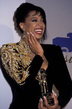 a woman holding an award in her right hand and wearing a black dress with gold sequins on it