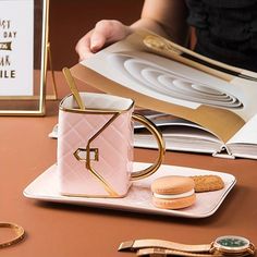 a pink coffee mug sitting on top of a white plate next to a cookie and a book