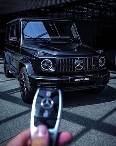 a hand holding a remote control in front of a black mercedes benz g - class