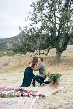 a man kneeling down next to a woman on top of a rug near a tree