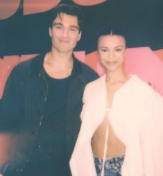 a man and woman standing next to each other in front of a pink wall with letters on it