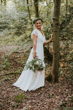 a woman standing in the woods wearing a white dress and holding a flower crown on her head