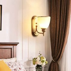 there is a lamp on the wall next to a bed and vase with flowers in it