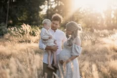 a man and woman holding a baby while standing in tall grass with the sun shining on them