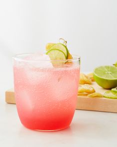 a pink drink in a glass with limes on the side and a cutting board behind it