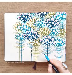 a hand holding a marker and drawing flowers on a notebook with colored pencils next to it