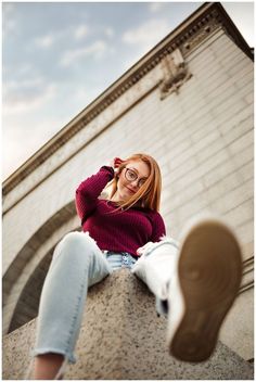 a woman sitting on top of a stone wall next to a brick building and wearing glasses